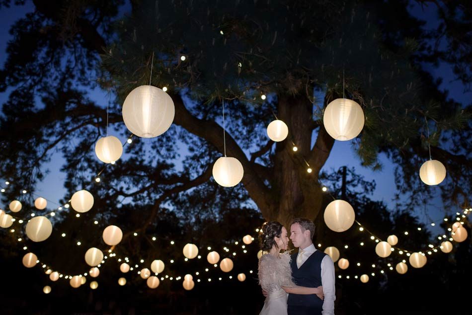 Image of a smartly dressed bride and groom embracing underneath a large tree with festoon lighting.